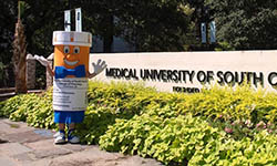college of pharmacy's pill bottle-shaped mascot stands outside and waves next to MUSC sign